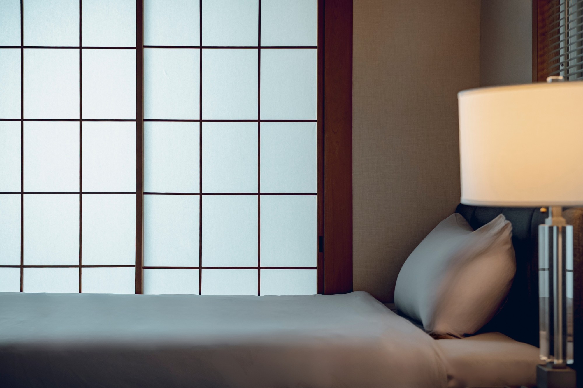 A bedroom in an ACOE property: a made bed beside a shoji screen, lit by a lamp.